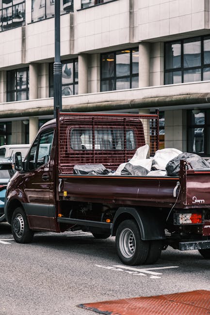 A small, dark red flatbed truck parked on an urban street in front of a modern office building with large glass windows and a concrete facade. The truck's cargo area is filled with various types of ru